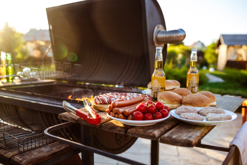 Luxury Outdoor Living A barbecue grill with sausages, burger patties, buns, cherry tomatoes, and two bottles of beer set on a wooden table outdoors—perfect for embracing luxury outdoor living. Toronto Contractors