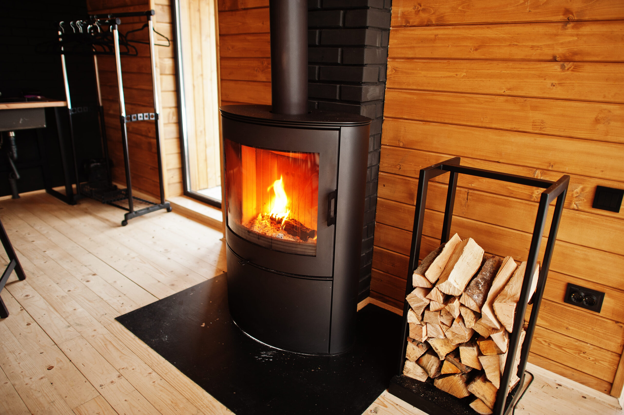 Luxury Outdoor Living A modern black wood stove with a fire burning inside stands on a black hearth in a wooden cabin, creating the perfect ambiance for luxury outdoor living. Nearby, a rack of firewood is stacked, ready to keep the warmth going. Toronto Contractors