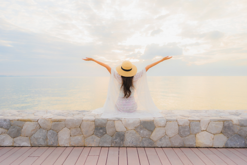 Luxury Outdoor Living A woman wearing a hat sits on a stone wall facing the sea with arms raised, embracing the essence of Luxury Outdoor Living under a cloudy sky at sunrise or sunset. Toronto Contractors