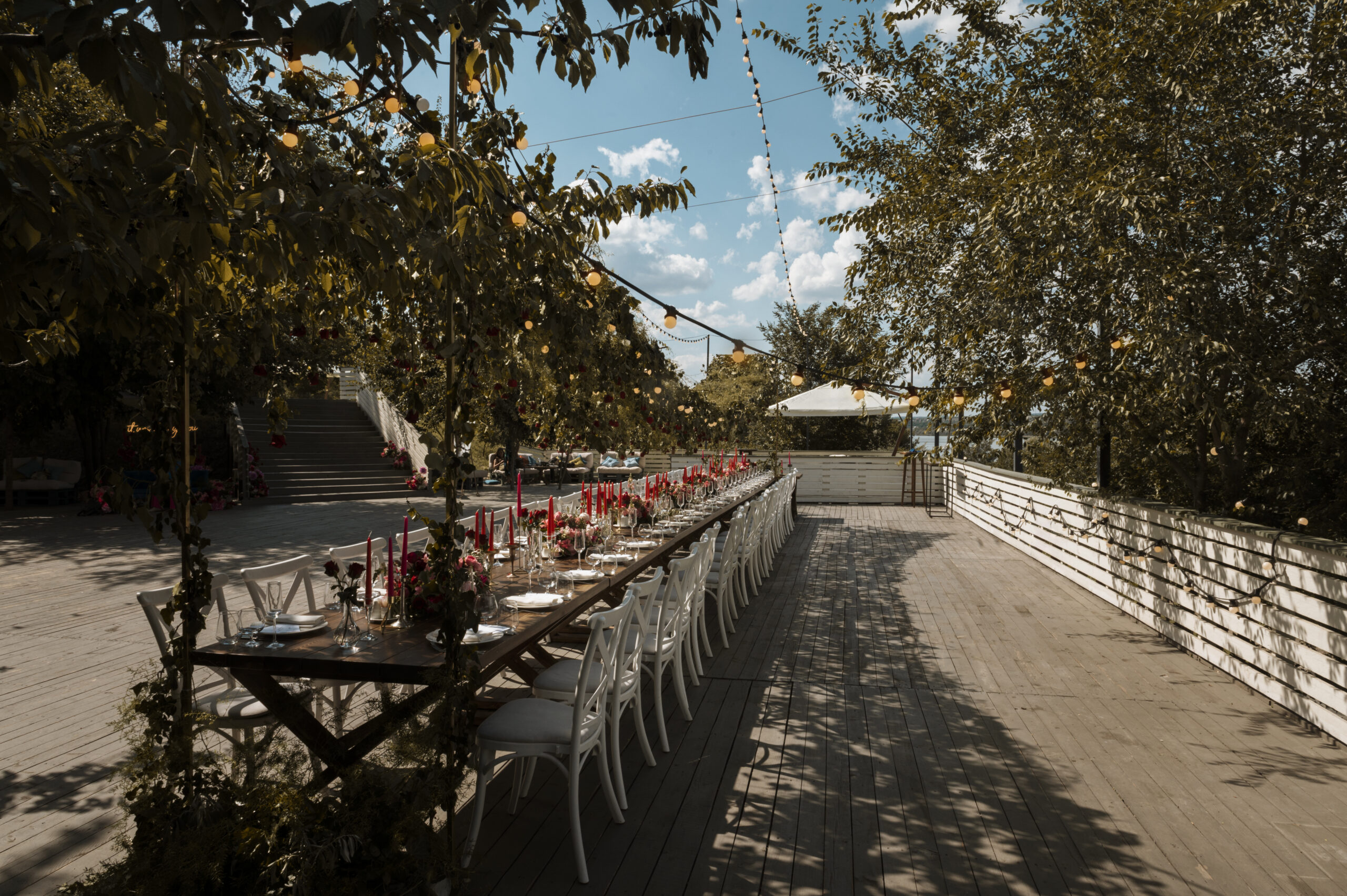 Luxury Outdoor Living A long outdoor dining table with white chairs is set with red flowers and candles on a wooden deck under string lights and trees, creating a Luxury Outdoor Living experience on a sunny day. Toronto Contractors