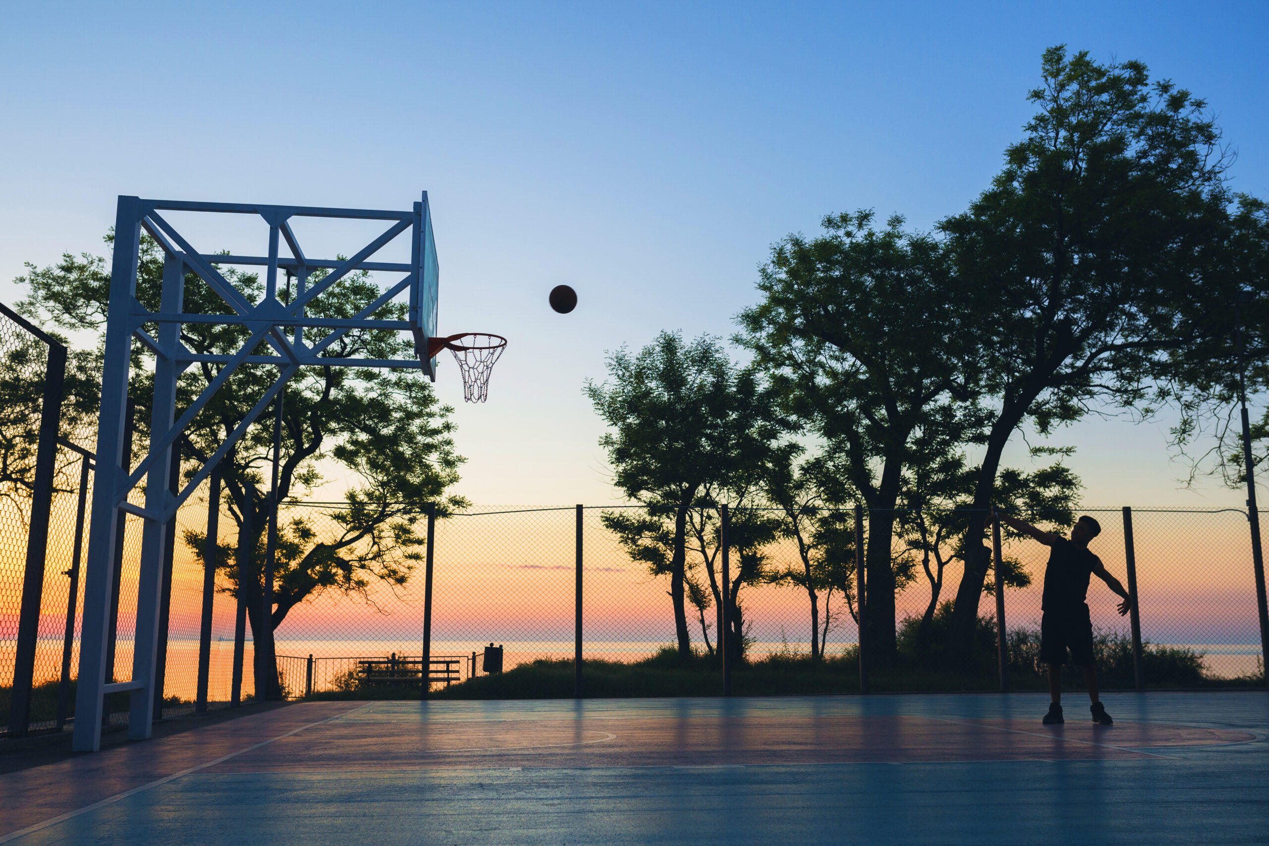 Luxury Outdoor Living A person shoots a basketball towards a hoop on an outdoor court at sunset, enjoying luxury outdoor living with trees and a body of water in the background. Toronto Contractors