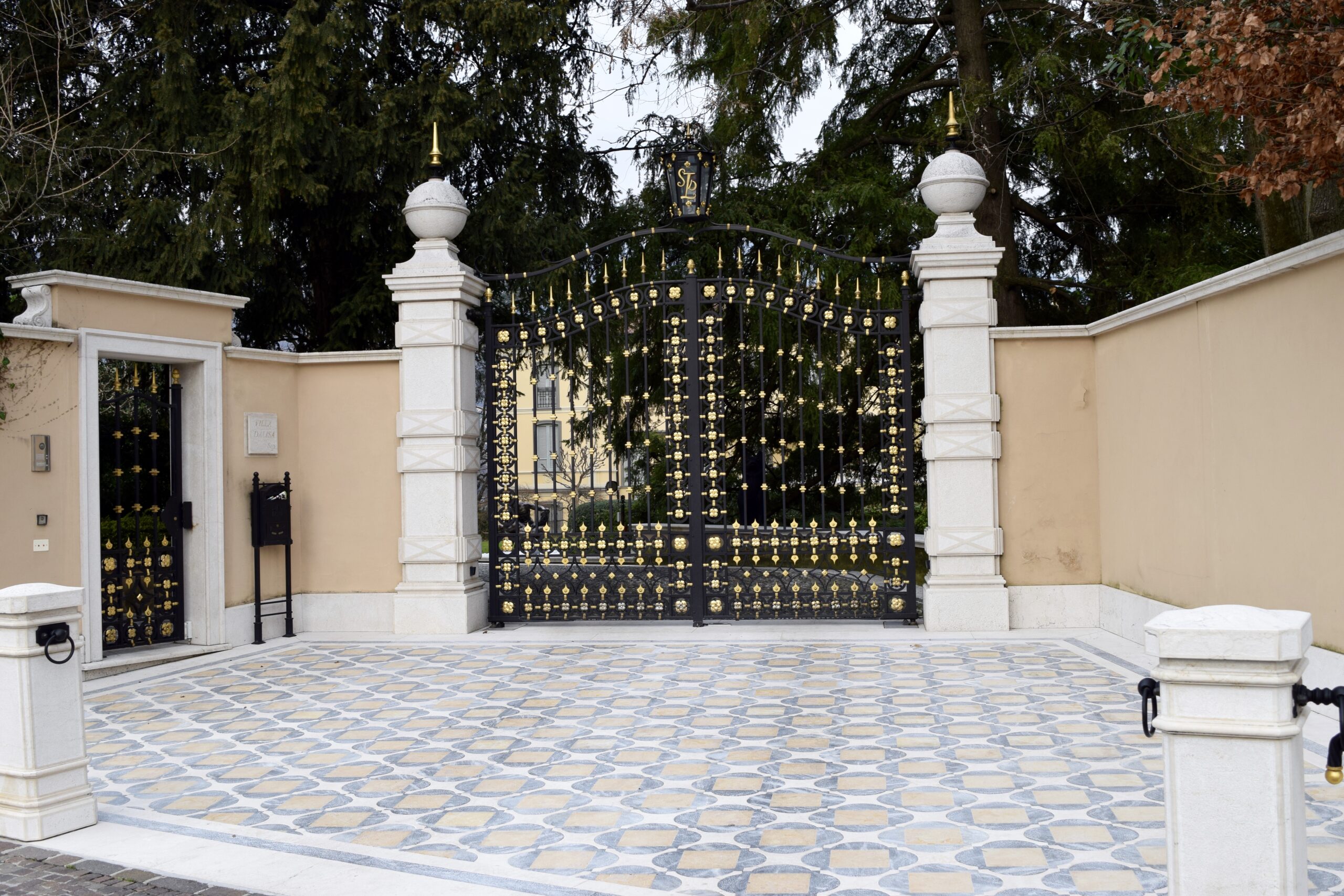 Luxury Outdoor Living Ornate black and gold gate with white stone pillars and beige walls, set on a patterned tile driveway, surrounded by trees—an elegant entrance that defines luxury outdoor living. Toronto Contractors