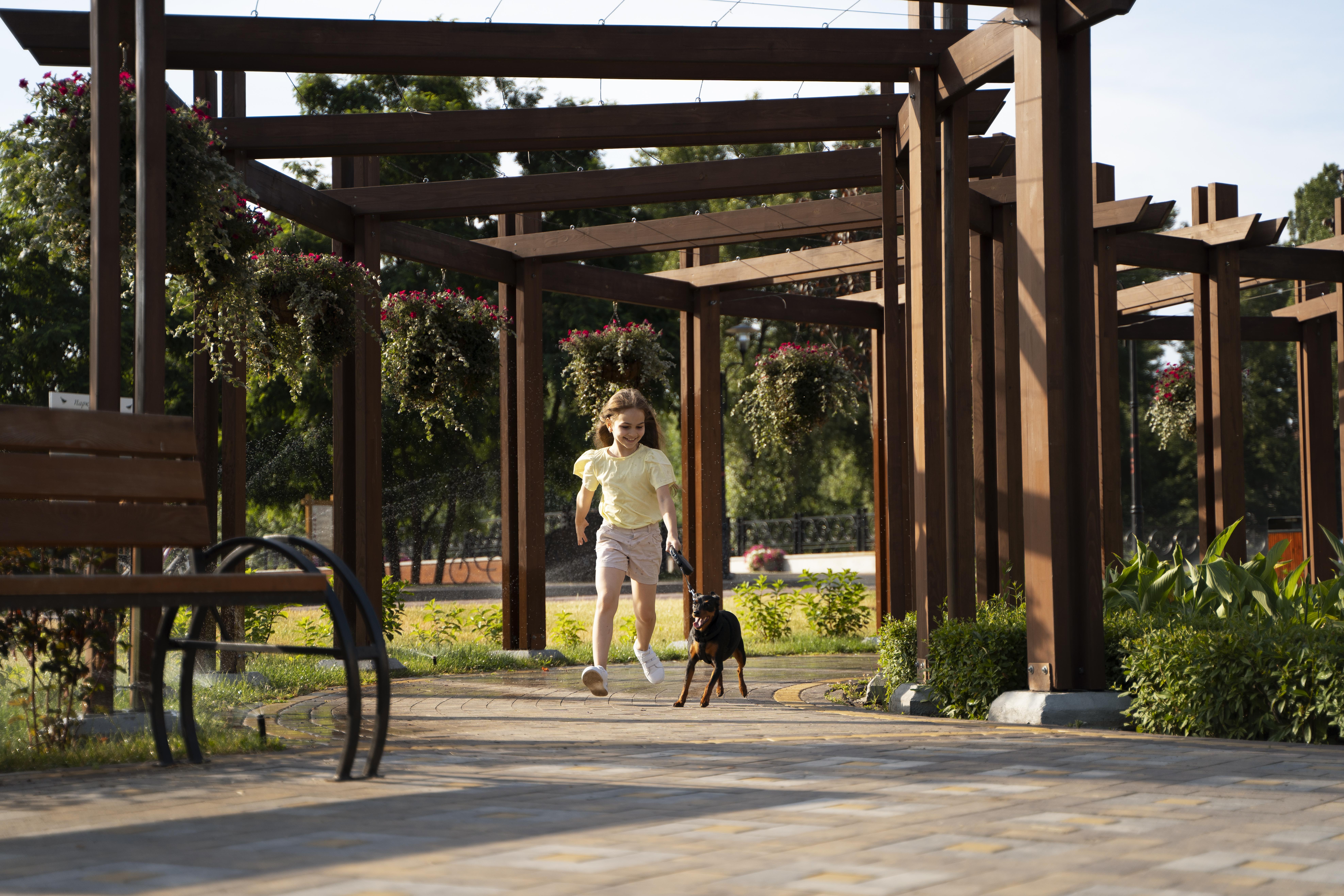 Luxury Outdoor Living A young girl in a light yellow shirt and shorts runs with a small black and brown dog on a leash under a wooden pergola, showcasing the joys of luxury outdoor living in the park. Toronto Contractors