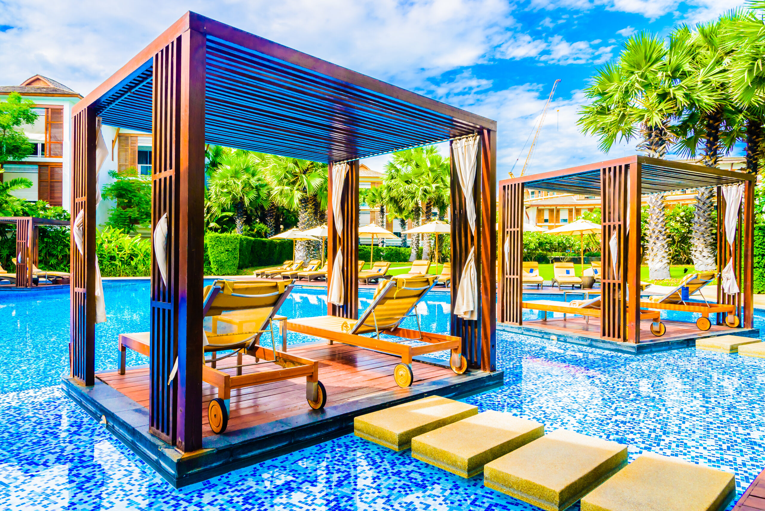Luxury Outdoor Living Experience luxury outdoor living in wooden poolside cabanas with lounge chairs, set on platforms over blue-tiled water and surrounded by palm trees and lush greenery under a partly cloudy sky. Toronto Contractors