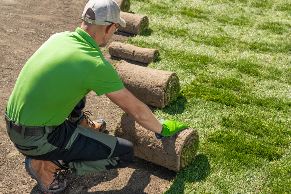 Luxury Outdoor Living A worker in green clothing and gloves is unrolling sod on a lawn, installing grass on a bare area to create a lush foundation for luxury outdoor living. Toronto Contractors