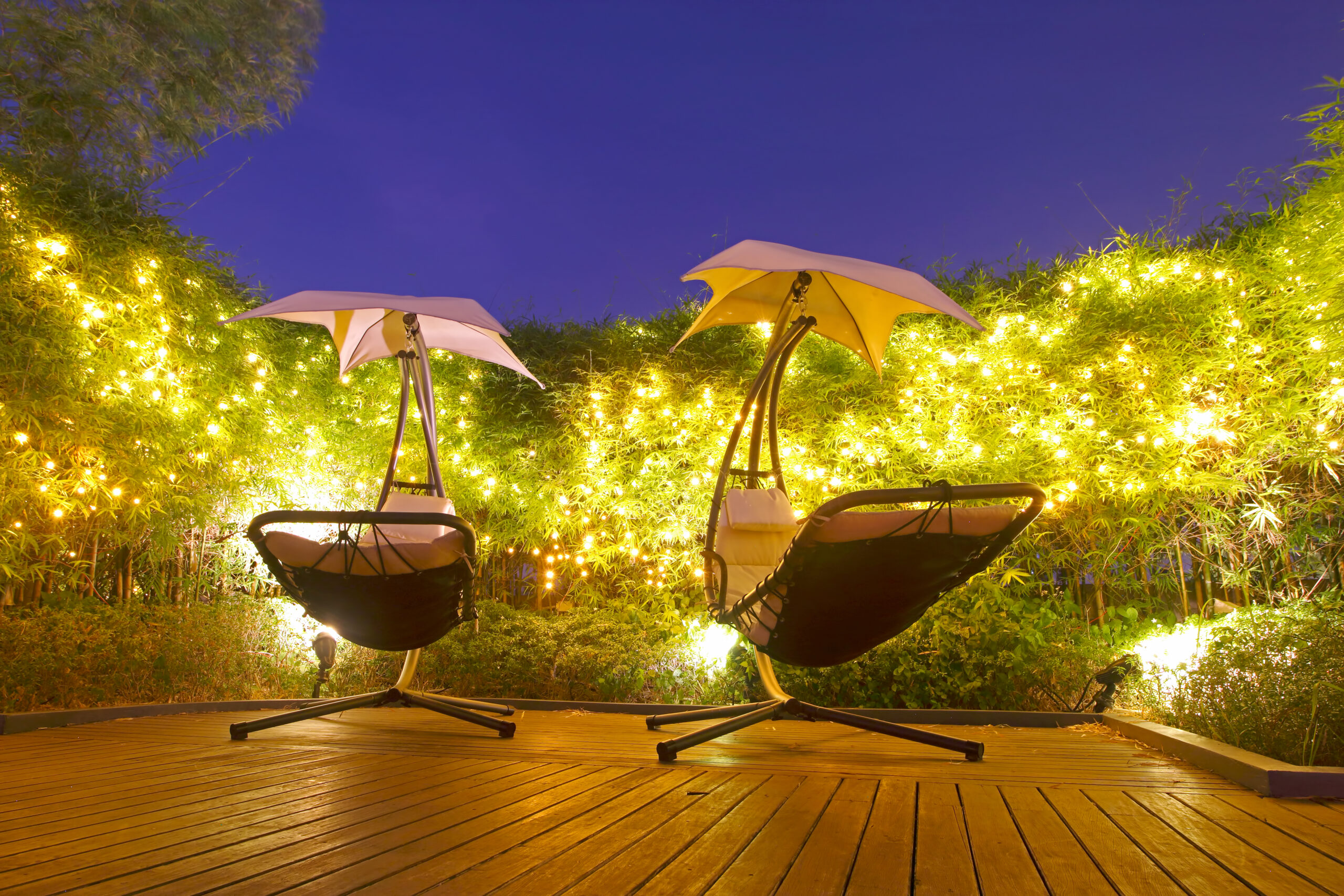 Luxury Outdoor Living Two lounge chairs with built-in umbrellas sit on a wooden deck, surrounded by illuminated greenery and string lights at dusk, creating a Luxury Outdoor Living experience. Toronto Contractors