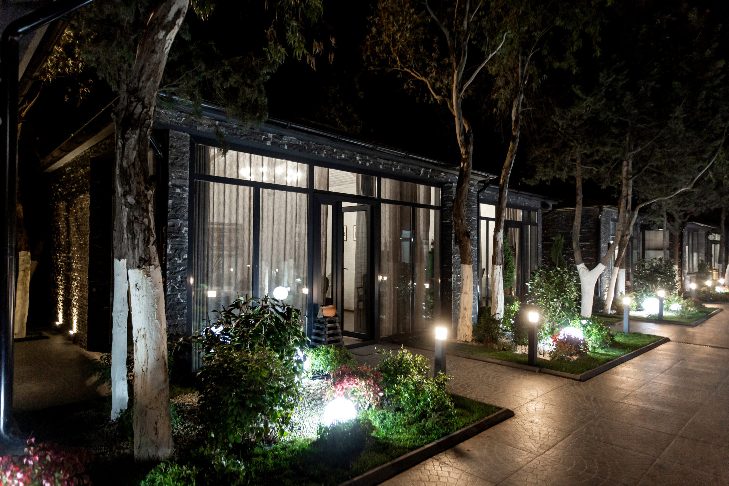 Luxury Outdoor Living Modern glass-fronted building with garden landscaping and illuminated pathway lights, photographed at night. Luxury Outdoor Living is showcased as trees line the walkway, creating an inviting and stylish atmosphere. Toronto Contractors