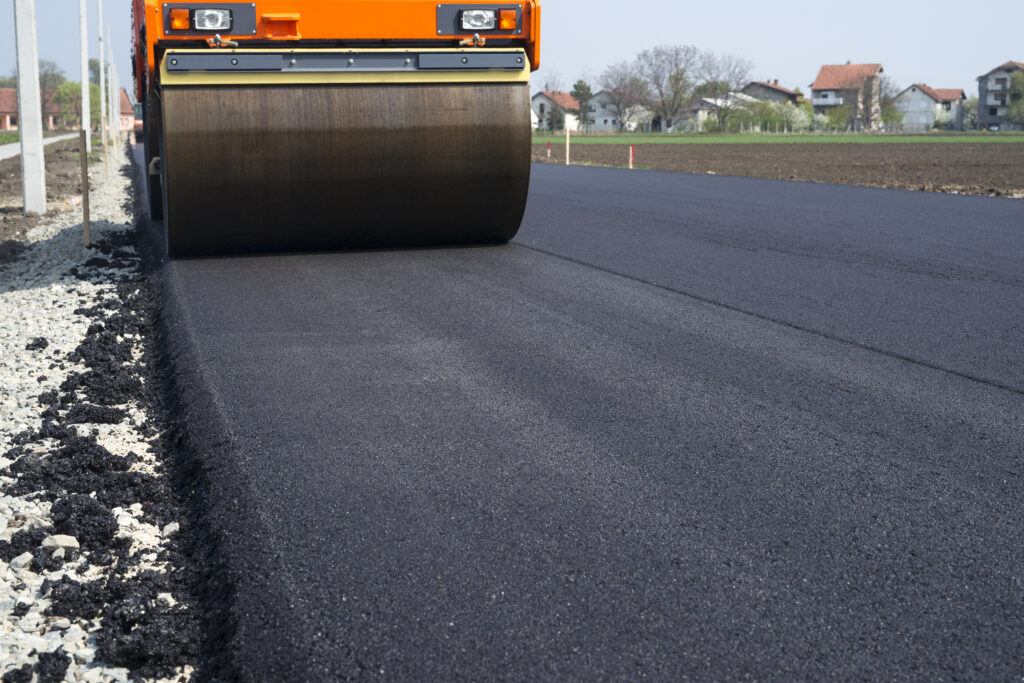 Luxury Outdoor Living A road roller compacts freshly laid asphalt on a new road, with rural houses and fields in the background—hinting at the promise of seamless access to luxury outdoor living. Toronto Contractors