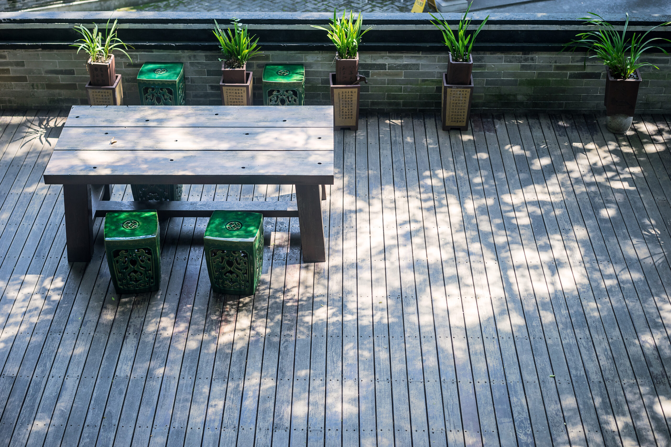 Luxury Outdoor Living Experience luxury outdoor living on a wooden deck featuring a rectangular table, four green ceramic stools, and lush potted plants against a brick wall. Sunlight and shadows create an inviting, serene retreat. Toronto Contractors