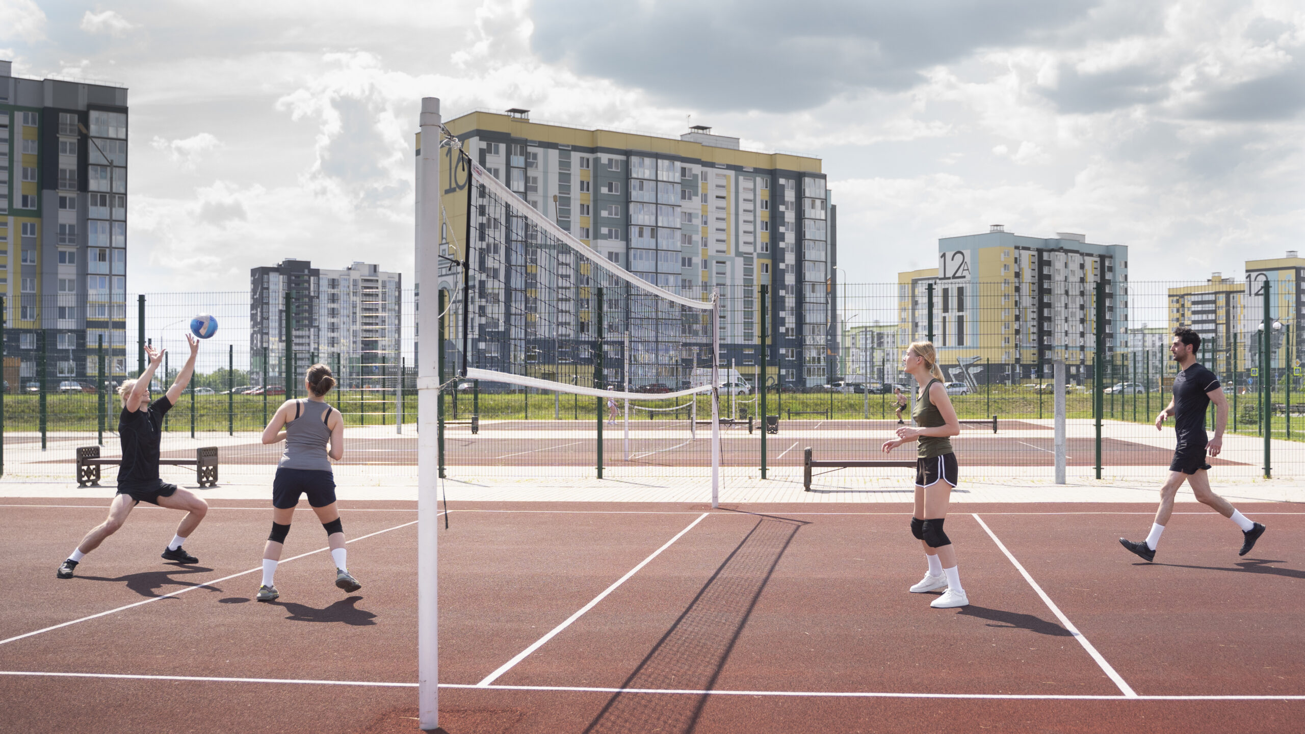 Luxury Outdoor Living Four people enjoy a game of volleyball on an outdoor court in an urban area, surrounded by apartment buildings and a partly cloudy sky—a perfect scene for luxury outdoor living. Toronto Contractors