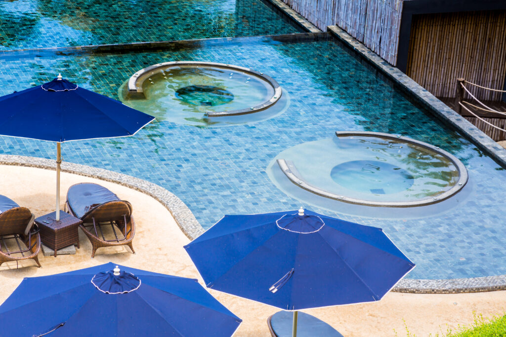 Luxury Outdoor Living A pool with two circular in-water seating areas offers Luxury Outdoor Living, surrounded by blue umbrellas and outdoor wicker chairs on a sandy patio. Toronto Contractors