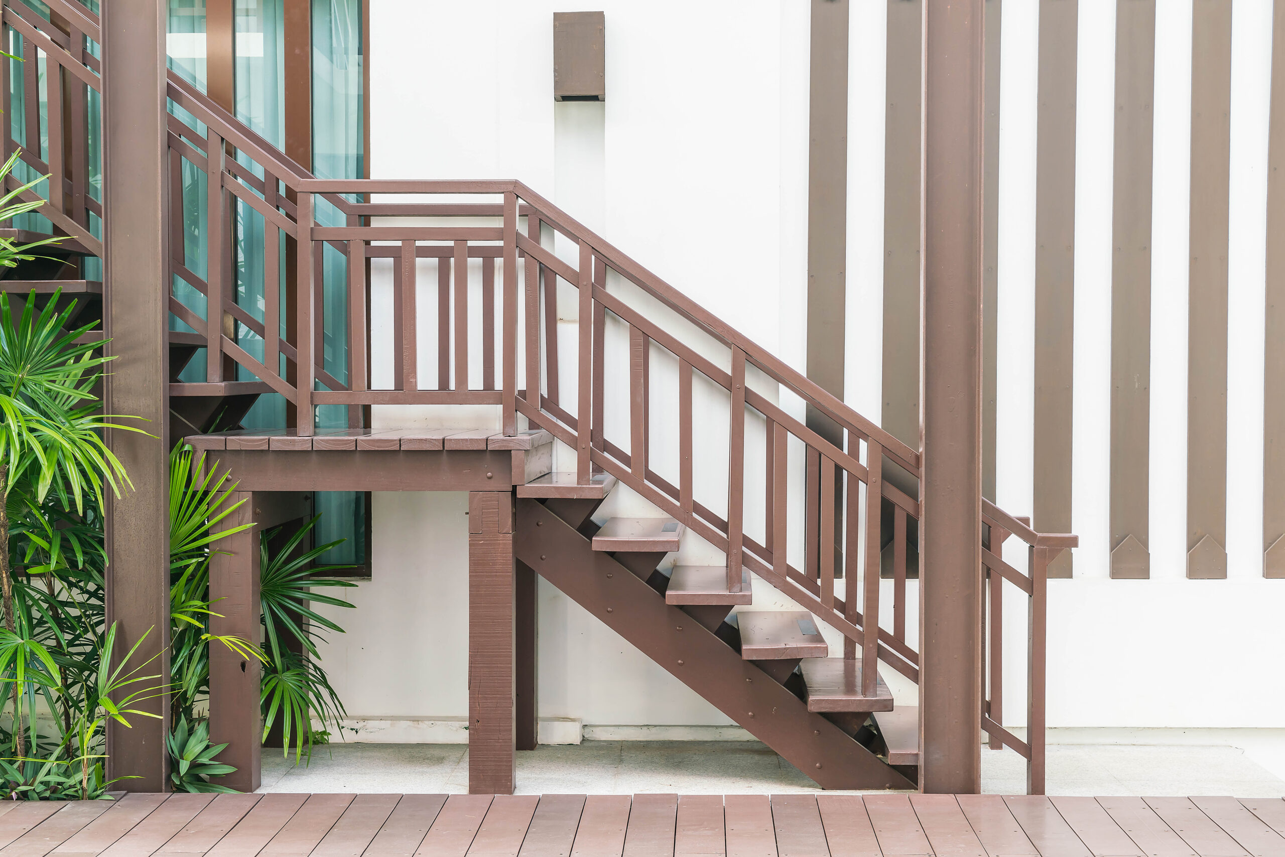 Luxury Outdoor Living Brown wooden staircase with railings beside a white building wall, surrounded by green plants and a wooden deck—perfectly blending luxury outdoor living into every detail. Toronto Contractors