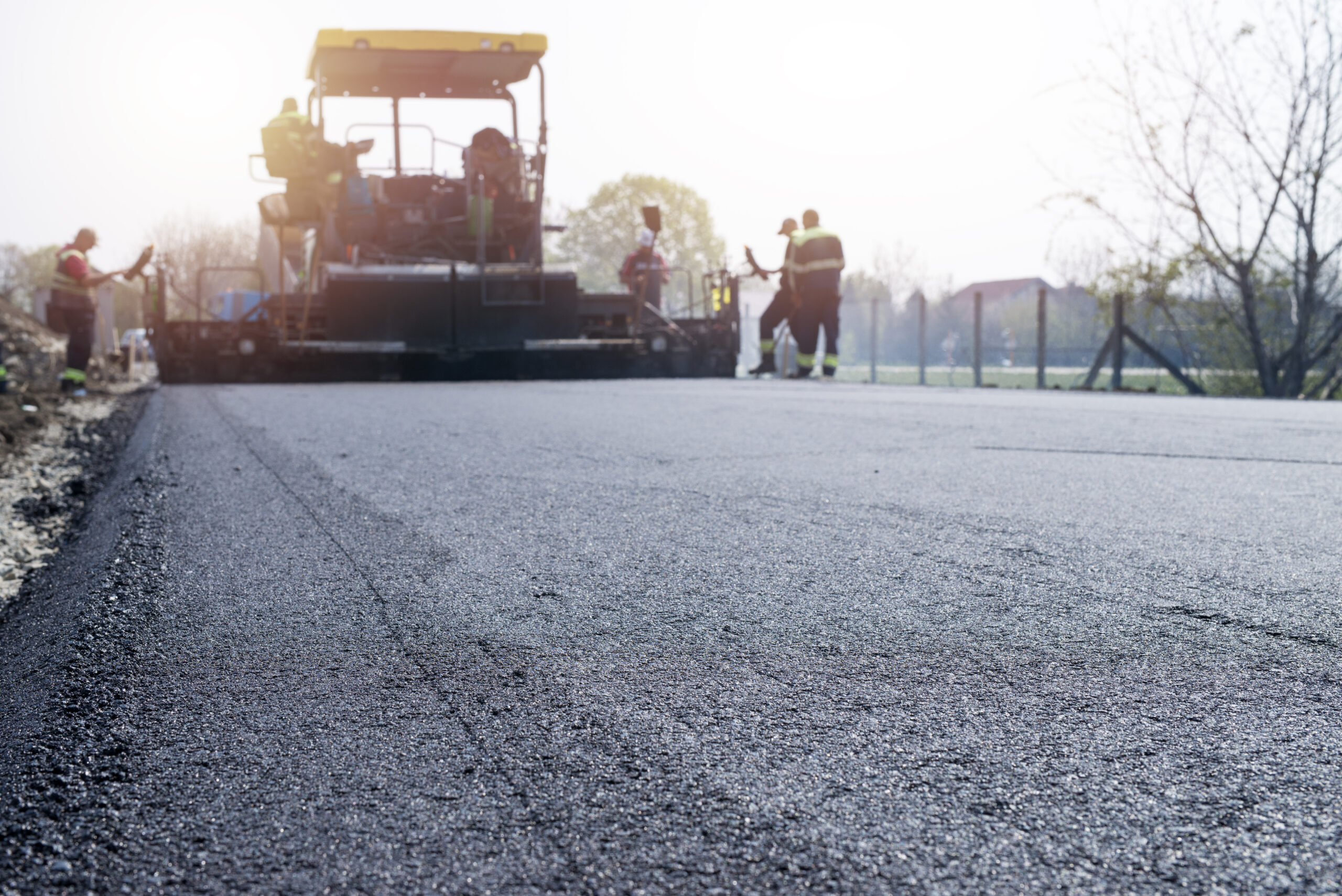 Luxury Outdoor Living Road construction workers use heavy machinery to lay new asphalt on a roadway in daylight, improving access for communities and future Luxury Outdoor Living developments, with some workers standing near the freshly paved surface. Toronto Contractors