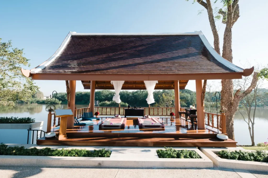 Luxury Outdoor Living Open-air wooden pavilion with a sloped roof, featuring massage beds, overlooks a calm river and lush greenery under a clear blue sky. Toronto Contractors