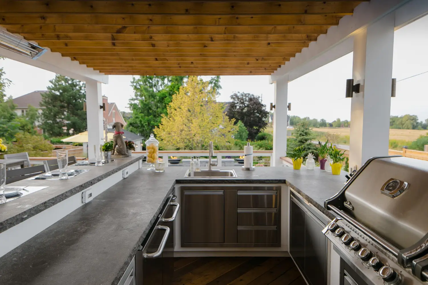 Luxury Outdoor Living Outdoor kitchen with stainless steel appliances, a built-in grill, sink, and bar seating under a wooden pergola, overlooking a landscaped backyard and trees. Toronto Contractors