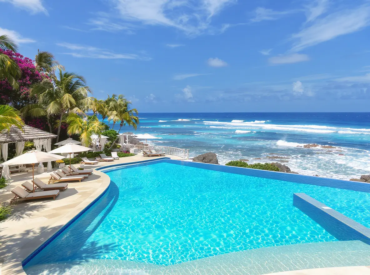Luxury Outdoor Living Infinity pool with lounge chairs and umbrellas overlooks a tropical beach and ocean under a blue sky with scattered clouds. Palm trees and flowers are visible on the left. Toronto Contractors