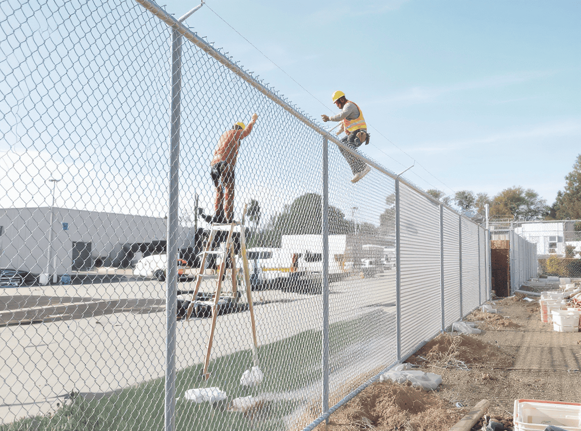 Luxury Outdoor Living Two construction workers in safety gear install or repair a chain-link fence; one stands on a ladder while the other works at the top of the fence. Toronto Contractors